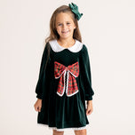 A young girl smiling and wearing a dark green velvet dress with a white Peter Pan collar, a red tartan bow at the waist, and a matching green bow in her hair, set against a plain light beige background.