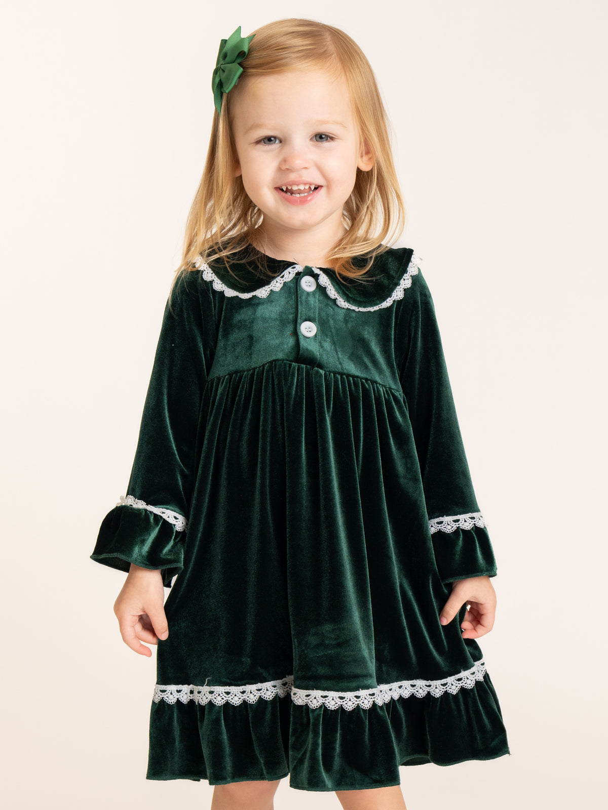A young girl with blonde hair wearing a dark green velvet nightgown with white lace trim and a matching green hair bow, standing against a plain off-white background.