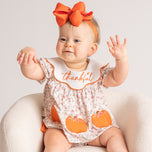 A baby wearing a short-sleeve floral romper with orange pumpkin appliques and a white bib that says 'thankful' in orange script, sitting in a cream-colored armchair. The baby is also wearing a large orange bow headband.
