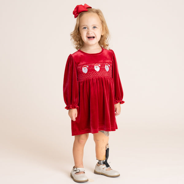 A young child wearing a festive red velvet dress with embroidered Santa faces across the chest, a matching red bow in her hair, and a prosthetic leg, standing against a plain beige studio background.