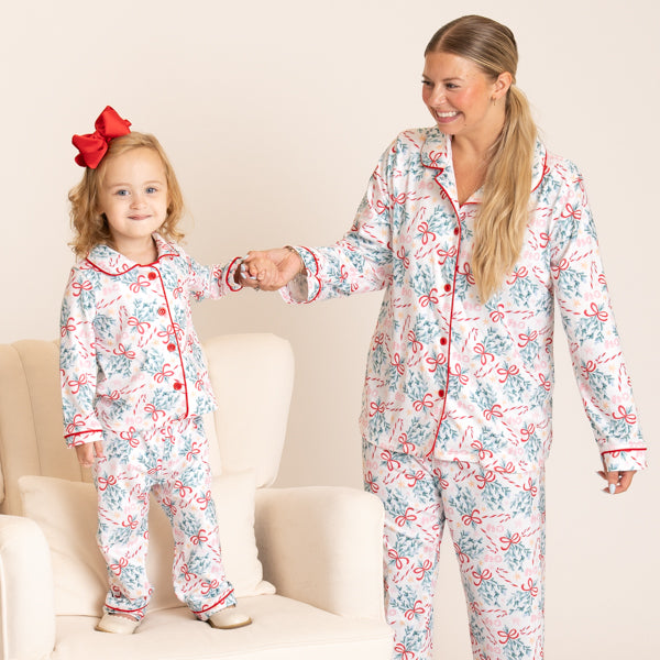 A woman and a young girl wearing matching holiday-themed pajamas with a white base, red trim, and a print of red bows and blue ornaments, standing in a living room setting.