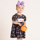 A toddler girl wearing a black short-sleeve dress with a purple and orange floral pattern, a white smocked chest panel with a pumpkin garland, and a large purple bow headband, holding an orange pumpkin bucket against a plain off-white background.