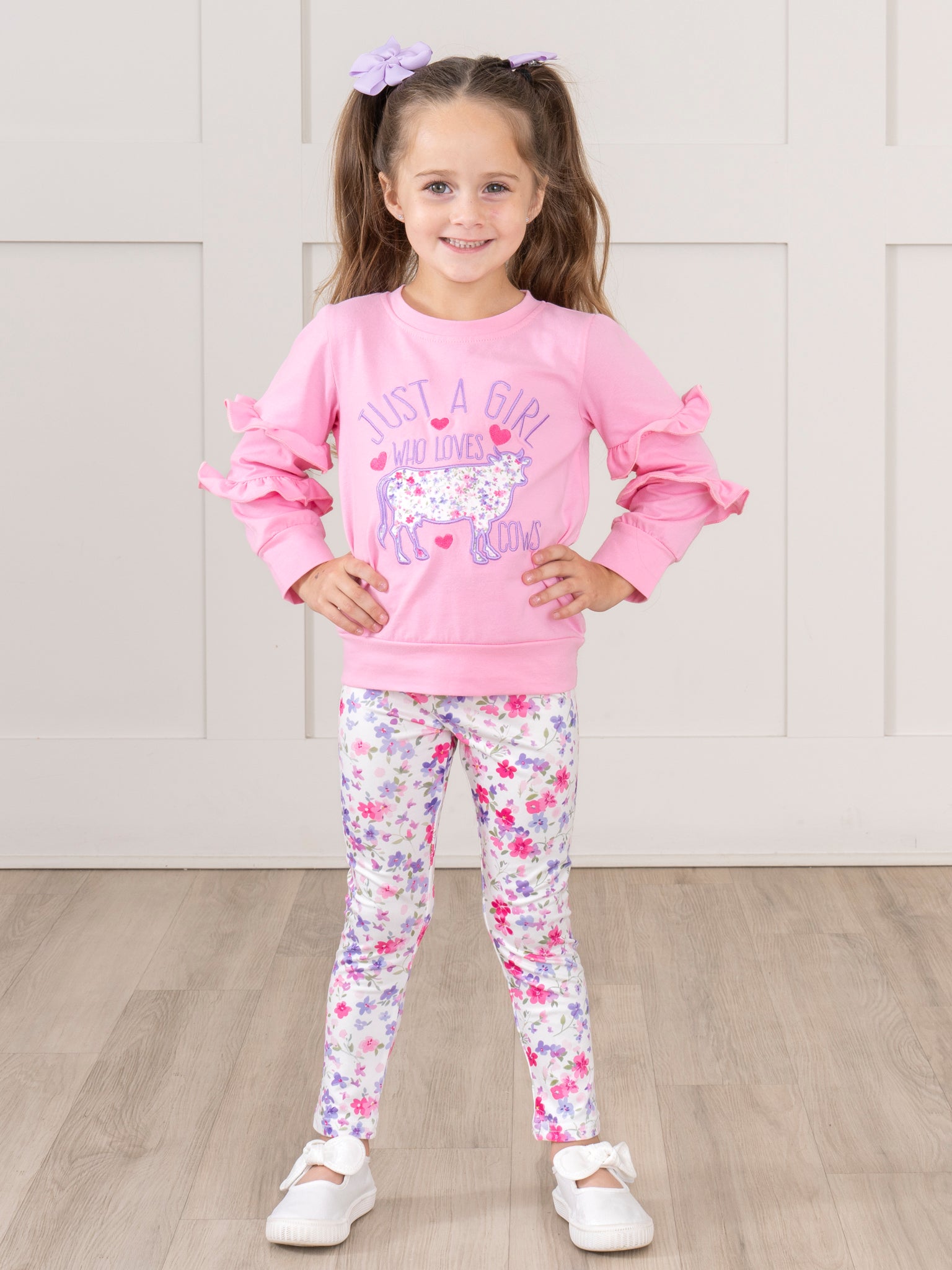 Young girl wearing a pink sweatshirt and floral leggings on a wooden floor.