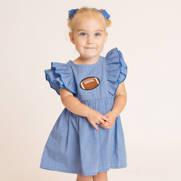 A young toddler girl with blonde hair in blue bows is wearing a light blue pinstriped dress with ruffled sleeves and a brown football applique on the chest, standing against a plain beige studio background.