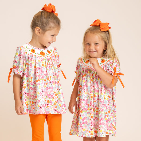 Two young girls modeling matching short-sleeve floral dresses with orange bow hair accessories, standing against a plain beige studio background.