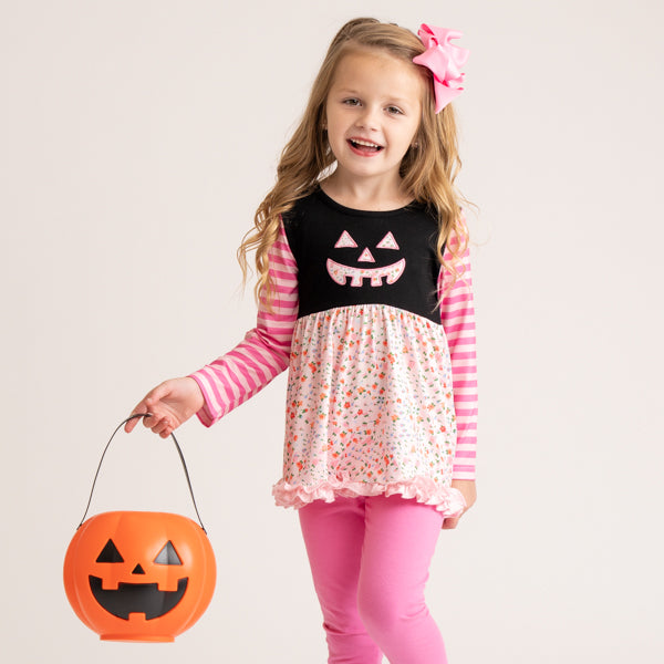 A young girl with a pink bow in her hair is wearing a Halloween costume dress with a black top featuring a jack-o'-lantern face, pink and white striped sleeves, and a white skirt with a small orange pattern. She is holding an orange pumpkin-shaped plastic bucket.