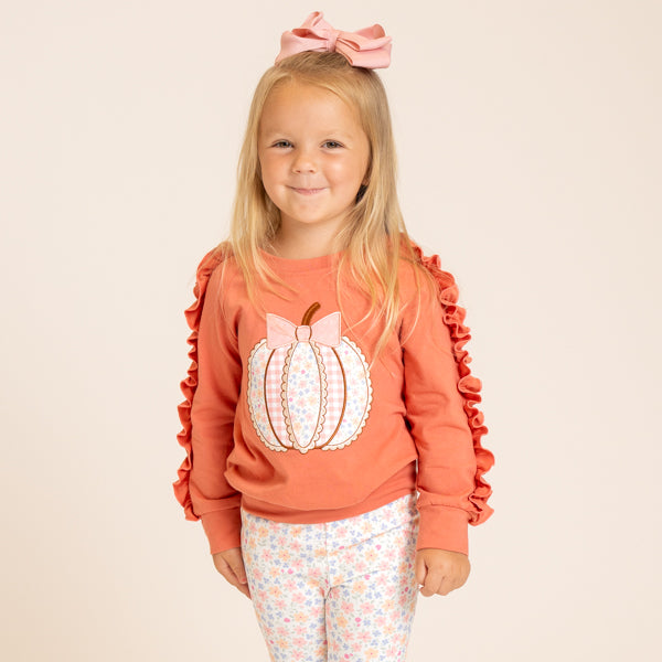 A young girl with blonde hair wearing a pink bow headband, a peach-orange long-sleeve top with ruffled sleeves and a pumpkin applique, and white patterned leggings, standing against a solid light beige background.