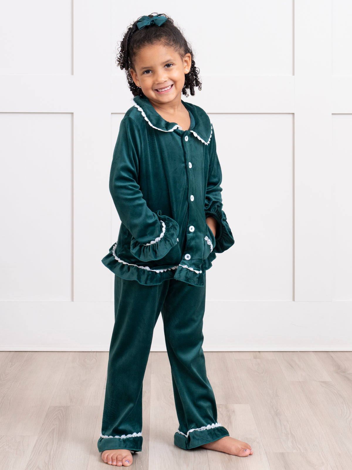 Young girl wearing a green velvet pajama set with white trim against a white paneled wall background