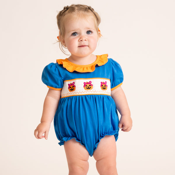 A toddler girl wearing a bright blue romper with orange ruffle trim, a white smocked chest panel with embroidered jack-o'-lanterns, and pink bows, photographed against a plain light beige studio background.
