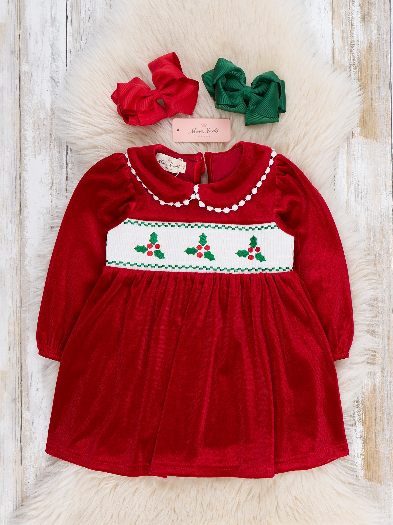 A red velvet long-sleeve dress with a white smocked chest panel featuring green holly and red berries, styled with a red bow and a green bow on a white faux fur rug.