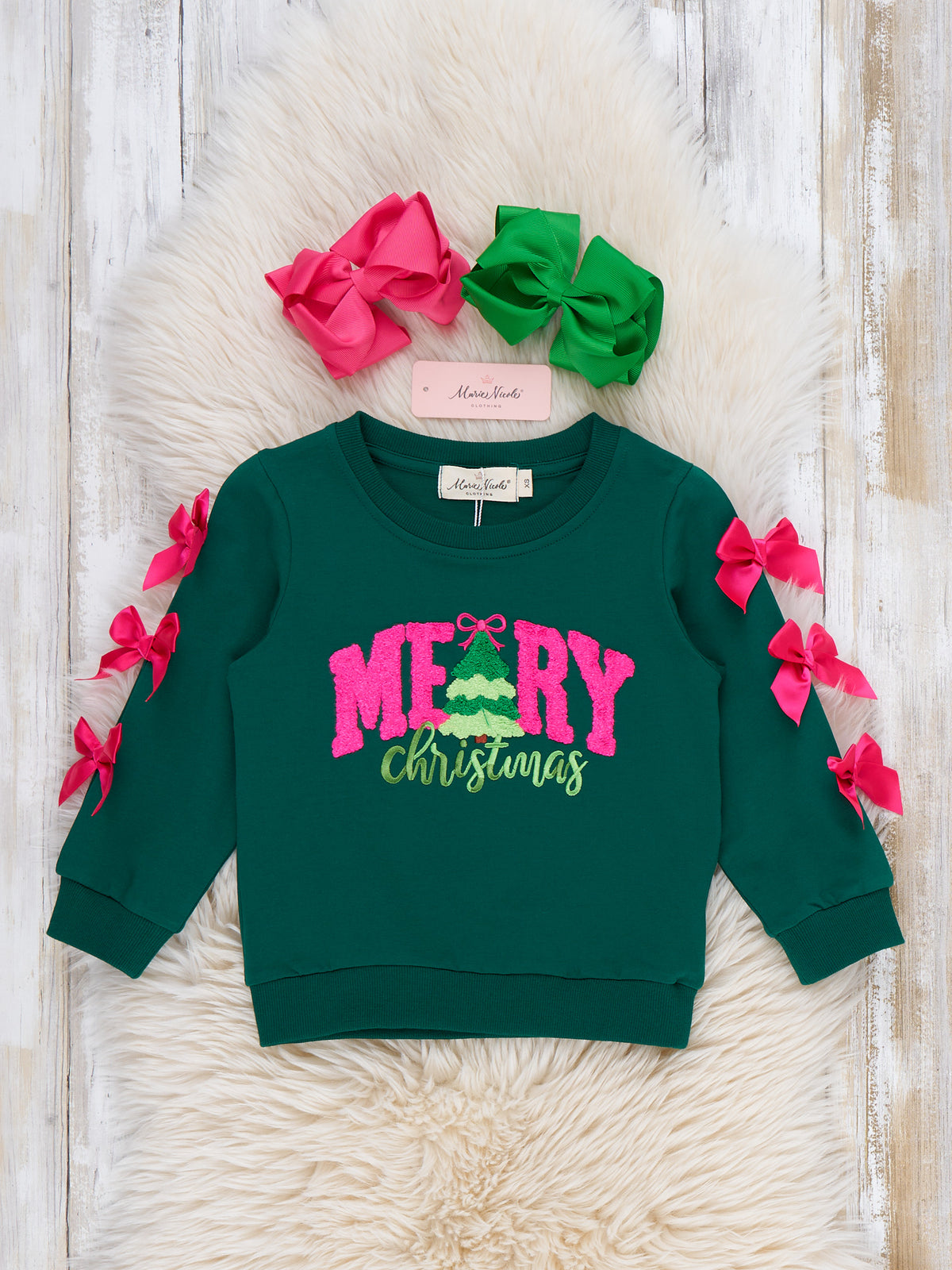 A toddler's dark green crewneck sweatshirt with 'MERRY Christmas' printed on the front, decorated with pink and green bows on the sleeves. The sweatshirt is displayed on a white faux fur rug with two matching hair bows and a brand tag.