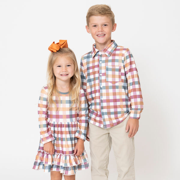 A young boy and girl modeling matching plaid outfits against a plain white studio background. The girl wears a long-sleeve ruffled dress and the boy wears a matching button-up shirt, both in a muted plaid pattern of beige, pink, blue, and white, with the girl wearing a brown bow in her hair.