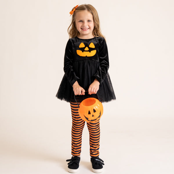 A young girl modeling a black velvet long-sleeve dress with a yellow-orange jack-o'-lantern face on the chest, paired with orange and black striped tights, holding a pumpkin-shaped bucket against a plain light beige studio background.