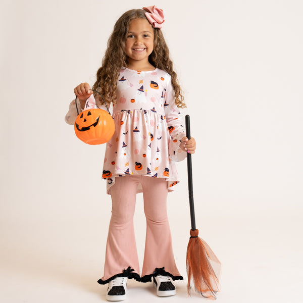 A young girl modeling a pink and white Halloween costume set, featuring a long-sleeve top with a pumpkin and witch pattern, matching pink bell-bottom pants, and black-and-white sneakers. She holds an orange jack-o'-lantern bucket and a broomstick against a solid light beige studio background.